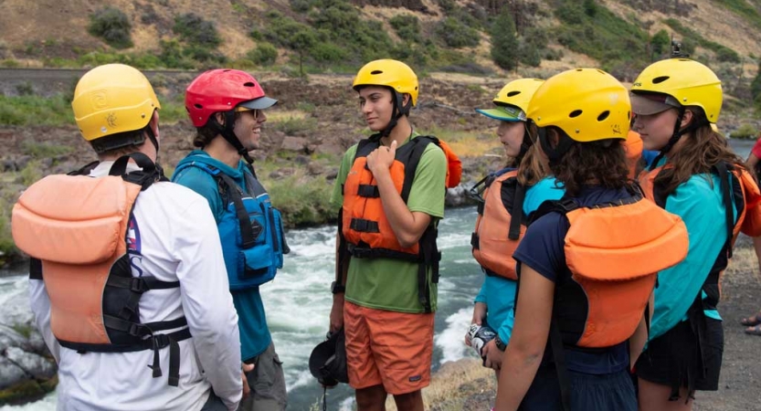 a group of outward bound students wearing helmets and life jackets talk to each other while standing above a river